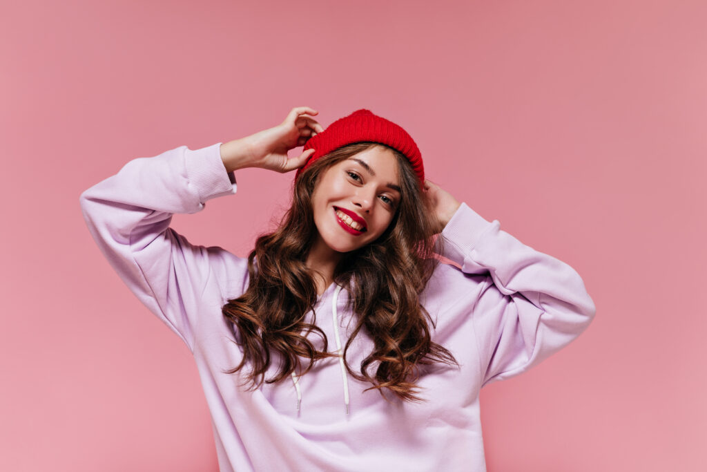 Charming cool girl in purple hoodie puts on red hat. Pretty brunette curly woman smiles widely on pink isolated background.