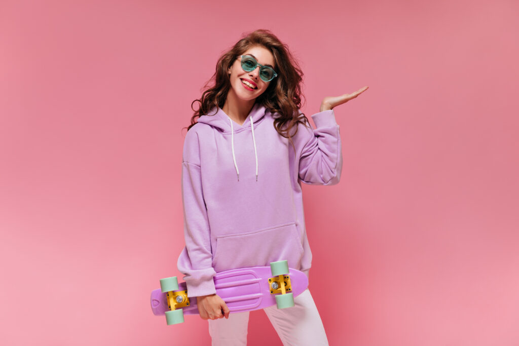 Charming girl in purple hoodie and white pants smiles and holds colorful longboard. Happy woman in green sunglasses poses on pink isolated background.