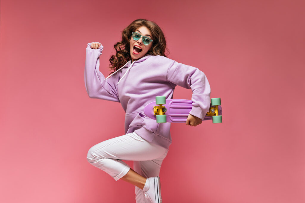 Cheerful cool girl in purple hoodie and white pants jumps on isolated. Happy brunette woman in green sunglasses holds colorful longboard on isolated pink background.