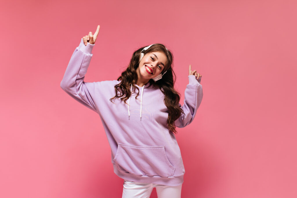 Happy cool girl in purple oversized hoodie points up and smiles widely. Charming brunette girl in purple outfit listens to music in headphones and dances on pink background.