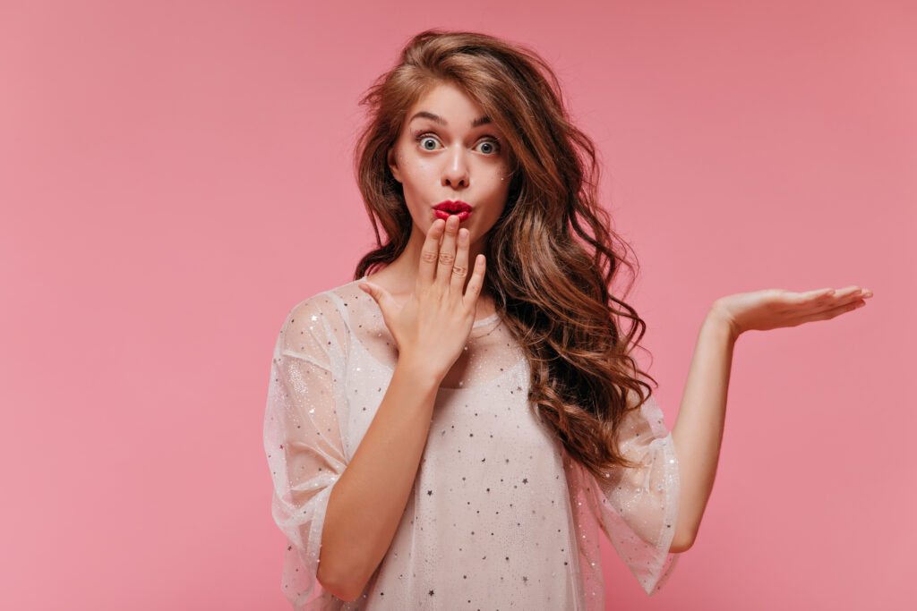 Portrait of curly long-haired woman in white stylish dress looking surprised. Shocked happy girl poses on pink isolated background.