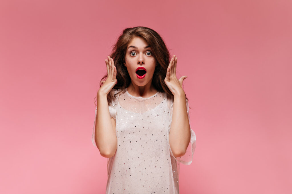 Portrait of shocked woman looking into camera. Charming brunette curly girl in white dress poses on pink isolated background.